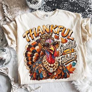 May include: A cream-colored t-shirt featuring a graphic of a turkey wearing leopard print glasses, a matching headband, and a pearl necklace. The text reads THANKFUL Grateful and Fabulous, surrounded by orange and yellow autumn leaves.