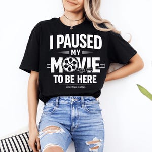 Op de afbeelding: Zwart T-shirt met de witte tekst "I PAUSED MY MOVIE TO BE HERE". Het woord "MOVIE" heeft een filmrol-afbeelding. De zin "priorities matter" staat eronder gedrukt.