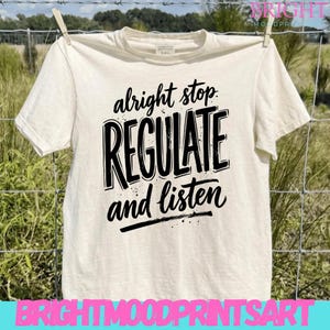 May include: Cream-colored t-shirt with the words "alright stop REGULATE and listen" in black, handwritten-style font. The shirt is hanging on a clothesline with a wooden clothespin. The background features a grassy field and a wire fence.