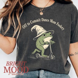 May include: Dark gray t-shirt featuring a green frog wearing a wizard hat and riding a scooter. The text "Off to Commit Dance Mom Foolery" arches across the top. The bottom left corner has the text "BRIGHT MOOD Prints Art".
