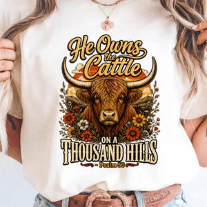 May include: A cream-colored t-shirt featuring a graphic of a brown highland cow with long horns, surrounded by orange and white flowers. The text reads "He Owns the Cattle ON A THOUSAND HILLS - Psalm 50" in a stylized, vintage-inspired font.