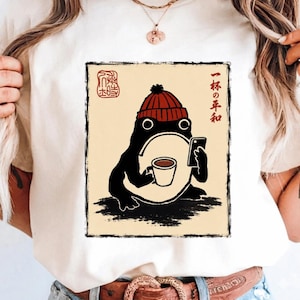 May include: A cream-colored t-shirt featuring a graphic of a black frog wearing a red beanie, holding a white coffee mug and a black smartphone. The design includes Japanese text and a red stamp-style seal on a light beige rectangular background.