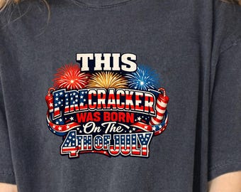 Op de afbeelding: Een antracietgrijs t-shirt met een afbeelding van rood, wit en blauw vuurwerk en de tekst THIS FIRECRACKER WAS BORN On The 4TH OF JULY. Het ontwerp bevat sterren- en strepenpatronen, met twee illustraties van vuurwerk aan de zijkanten.