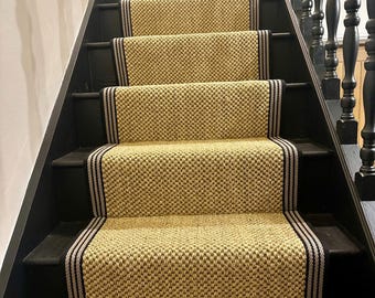 Puede incluir: Una escalera con escalones pintados de negro y alfombras tejidas beige. Cada alfombra tiene un borde negro con dos finas rayas claras. La escalera tiene una barandilla de madera oscura a la derecha y una pared blanca a la izquierda.