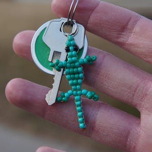 May include: A teal beaded lizard keychain with black eyes hangs from a silver key. The key has a green and silver fob. The keychain is held in a hand.
