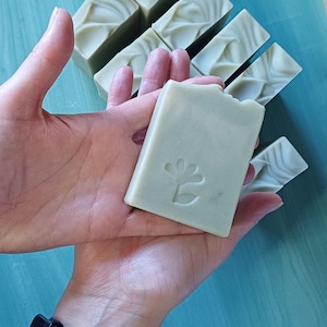 May include: Several light green rectangular bars of soap. One bar is held in a hand, displaying a flower design. The other bars have a textured pattern. The soap is a pale green color.