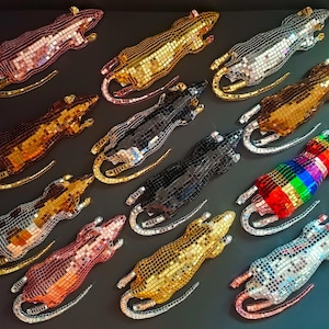 May include: A collection of twelve rat-shaped ornaments covered in small, reflective mirror tiles. The rats are arranged in various colors including gold, silver, black, copper, red, and a multi-colored rainbow pattern, each featuring a long tail.