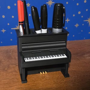 May include: A black piano-shaped desk organizer holding several pens and markers. The organizer has a detailed keyboard design with white and black keys. A red marker and several black markers are visible.