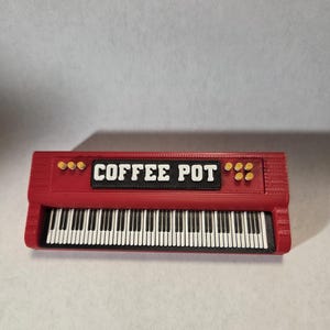 May include: A red rectangular object shaped like a keyboard with a black center panel featuring the white text COFFEE POT. The top section includes three yellow circular buttons on the left and four on the right, above a standard piano key layout.