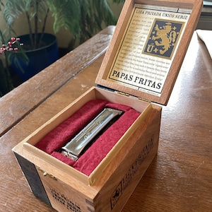 Puede incluir: Una caja de madera abierta con tapa con bisagras, forrada con tela roja, que contiene una armónica plateada. La tapa presenta texto y un logotipo. La caja está etiquetada como "LIGA PRIVADA UNICO SERIE" y "PAPAS FRITAS".
