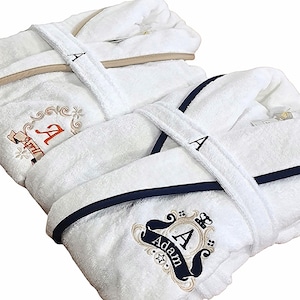May include: Two white terry cloth bathrobes with embroidered details. One robe has a tan trim and the other has a navy blue trim. Both robes feature the letter "A" embroidered on the chest and the sash. One robe has the name "April" embroidered on it.
