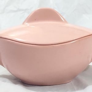 May include: A solid light pink sugar bowl with a matching lid and two side handles. The bowl features a smooth, rounded body and a lid with a prominent, curved handle on top. The design is simple, featuring a uniform pastel pink finish throughout.