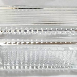 May include: A rectangular clear glass butter dish with a lid. The lid features a series of horizontal raised lines and a decorative beaded border. The base of the dish has vertical ribbed detailing along the sides, creating a textured appearance.