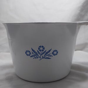 Könnte beinhalten: Weißer Emaille-Krug mit blauem Blumendesign. Der Krug hat einen Griff und einen ausgestellten Rand. Das blaue Blumendesign ist auf der Vorderseite des Kruges zentriert. Der Krug steht auf einer weißen Oberfläche.