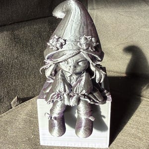 Puede incluir: Figura plateada de un gnomo sentado con un gorro puntiagudo adornado con flores. El gnomo lleva un vestido y botas, y está sentado sobre un bloque blanco. La figura tiene una estética caprichosa y de cuento de hadas.