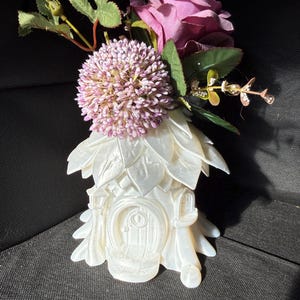 Könnte beinhalten: Eine weiße, märchenhafte dekorative Vase in Form eines Hauses, verziert mit blattartigen Details. Die Vase ist mit einem Cluster aus lila Blumen und grünem Laub bedeckt. Das Haus hat eine detaillierte Tür und Fenster.