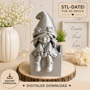 May include: A silver-colored 3D printed figurine of a small character with a large pointed hat adorned with flowers, sitting on the edge of a square box. The image includes text that reads STL-DATEI FÜR 3D DRUCK and SOFORT DOWNLOAD.