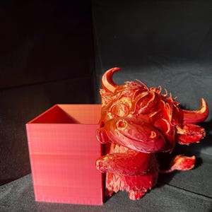 May include: A red 3D-printed Highland cow planter with a matching square planter. The cow has horns, a fluffy mane, and a smiling face. The planter is a solid, geometric shape, perfect for small plants or succulents.