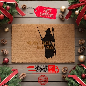 May include: A rectangular coir doormat with the text "SHOES SHALL NOT PASS" and a black silhouette of a wizard. The mat is surrounded by Christmas decorations, including red ribbons, ornaments, and pinecones.