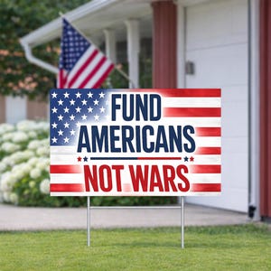 May include: A yard sign with the American flag design and the text "FUND AMERICANS NOT WARS" in red, white, and blue. The sign is displayed on a green lawn with a house and American flag in the background.