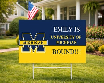 May include: A yard sign with a navy blue and yellow design. The sign features the University of Michigan logo and the text "EMILY IS UNIVERSITY OF MICHIGAN BOUND!!!" displayed on a green lawn with a house and American flag in the background.
