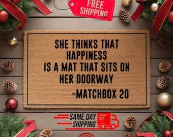 May include: A rectangular coir doormat with the text "SHE THINKS THAT HAPPINESS IS A MAT THAT SITS ON HER DOORWAY -MATCHBOX 20" in black. The mat is surrounded by Christmas decorations, including pine boughs, ornaments, and ribbons.