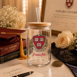 K&ouml;nnte beinhalten: Ein klares Glas mit einem Holzdeckel und einem Glasstrohhalm. Auf dem Glas ist das Wappen der Harvard University und der Name "Jennifer" in roter Schrift abgebildet. Eine Abschlusskappe, B&uuml;cher und eine Karte mit der Aufschrift "Herzlichen Gl&uuml;ckwunsch zum Abschluss!" sind ebenfalls zu sehen.