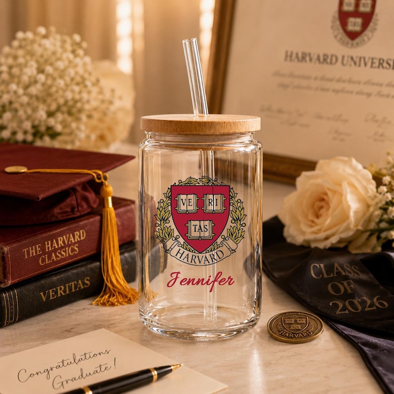 K&ouml;nnte beinhalten: Ein klares Glas mit einem Holzdeckel und einem Glasstrohhalm. Auf dem Glas ist das Wappen der Harvard University und der Name "Jennifer" in roter Schrift abgebildet. Eine Abschlusskappe, B&uuml;cher und eine Karte mit der Aufschrift "Herzlichen Gl&uuml;ckwunsch zum Abschluss!" sind ebenfalls zu sehen.