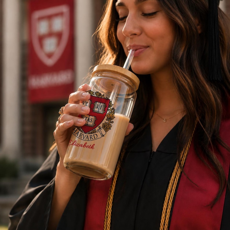 K&ouml;nnte beinhalten: Ein durchsichtiger Glasbecher mit Holzdeckel und Strohhalm, mit dem Harvard University-Wappen und dem Namen "Elizabeth" in roter Schrift. Der Becher ist mit einem hellfarbigen Getr&auml;nk gef&uuml;llt.