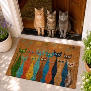 May include: A rectangular coir doormat featuring a colorful graphic print of ten stylized cats in various shades of green, orange, yellow, blue, and brown, accented by small star shapes, positioned on a concrete porch in front of a wooden door.