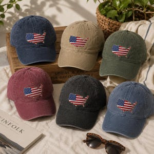 May include: A collection of six baseball caps in various colors including navy, tan, olive green, maroon, charcoal, and light blue. Each cap features an embroidered patch of the United States map filled with the American flag pattern on the front.