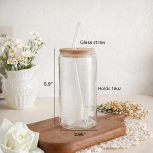 K&ouml;nnte beinhalten: Ein durchsichtiger Glasbecher mit Bambusdeckel und Glasstrohhalm. Der Becher ist 15 cm hoch und 7,5 cm breit und fasst 473 ml. Der Becher steht auf einem Holzschneidebrett, mit Blumen im Hintergrund.