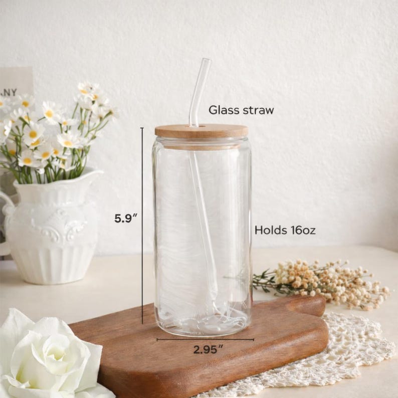 K&ouml;nnte beinhalten: Ein durchsichtiger Glasbecher mit Bambusdeckel und Glasstrohhalm. Der Becher ist 15 cm hoch und 7,5 cm breit und fasst 473 ml. Der Becher steht auf einem Holzschneidebrett, mit Blumen im Hintergrund.