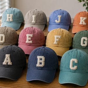 May include: A collection of eleven baseball caps in various colors, each featuring a large, cream-colored, embroidered capital letter on the front. The letters A through K are displayed across the caps, which have a washed, matte fabric finish.