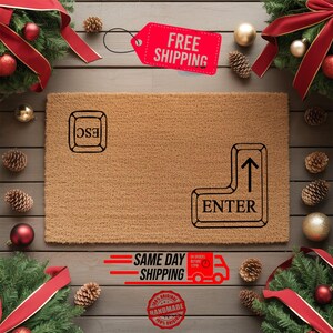 May include: A rectangular brown coir doormat with black text that reads "ESC" and "ENTER" in the shape of keyboard keys. The mat is surrounded by Christmas decorations, including red ribbons, ornaments, and pinecones.