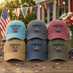 May include: Six baseball caps in various colors, including blue, gray, and red. Each cap features the text "AMERICA 250 WE THE FREE USA" with an eagle graphic. The caps are arranged on a wooden surface with patriotic decorations in the background.