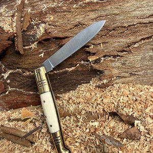 Könnte beinhalten: Ein Klappmesser mit polierter Stahlklinge und einem Griff aus cremefarbenem Material mit Messingakzenten. Das Messer ist geöffnet, die Klinge wird gezeigt, und liegt auf Holzspänen und einer strukturierten Holzoberfläche.