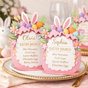 May include: Pink Easter brunch place cards with bunny ears, flowers, and colorful eggs. The cards read "Olivia" and "Sophia" with a menu of mini pancakes, fruit cups, carrot muffins, chocolate eggs, and bunny donuts.