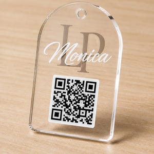 May include: Clear acrylic tag with rounded top and a hole for hanging. The tag features the name "Monica" in a script font, with the initials "LP" above. A black and white QR code is printed at the bottom.