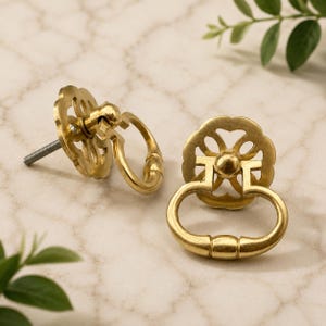 May include: Two brass drawer pulls featuring a circular backplate with a decorative cutout floral pattern and a hinged drop ring handle. Each piece includes a threaded metal bolt extending from the back for mounting to furniture surfaces.