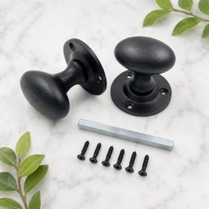 May include: Two matte black oval door knobs with circular mounting plates, each featuring three screw holes. The set includes a single square metal spindle bar and six matching black screws for installation, all presented as individual components.