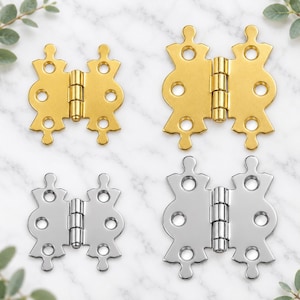May include: Four metal butterfly hinges arranged in a grid. Two hinges are gold-toned and two are silver-toned, with one of each colour being larger than the other. Each hinge features a central barrel and four circular screw holes on the wings.
