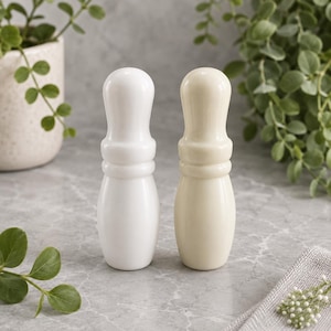 May include: Two smooth, glossy bowling pin-shaped objects standing upright. One object is a bright, stark white, while the other is a soft, creamy off-white. Both feature a rounded top, a narrow neck, and a wider, rounded base with a polished finish.