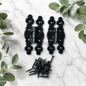May include: Two matte black metal hinges featuring an ornate, elongated shape with rounded ends and multiple pre-drilled screw holes. A small pile of matching black metal screws is positioned directly below the two hinges for installation.