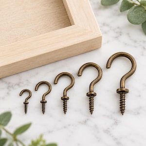 May include: A set of five antique brass-finished metal cup hooks arranged in increasing order of size. Each hook features a curved top and a threaded screw base, designed for installation into surfaces. The hooks have a metallic, weathered finish.