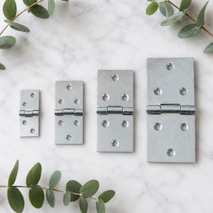 May include: Four rectangular silver-coloured metal hinges of varying sizes arranged in a row. Each hinge features a central barrel joint and multiple pre-drilled circular screw holes on both flat plates for mounting purposes.