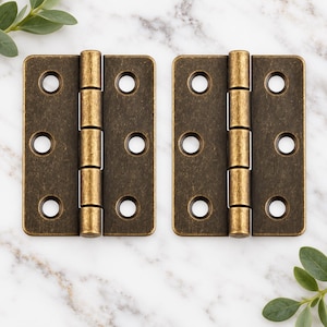 May include: Two rectangular metal door hinges with an antique brass finish. Each hinge features a central barrel with five knuckles and two flat plates, each containing three circular screw holes for mounting purposes.