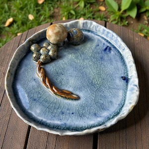 May include: A handcrafted ceramic plate with a unique design. The plate features a light blue, crackled glaze and an organic shape. It is adorned with a sculpted grape cluster and two spherical accents, adding a touch of nature-inspired artistry.