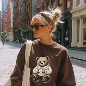 May include: A brown sweatshirt featuring a graphic of a teddy bear wearing a sweater and holding a cup of coffee. The bear is in a seated position. The sweatshirt is worn by a person wearing sunglasses and a leopard print headband.