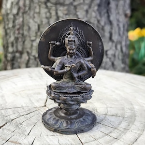 Könnte beinhalten: Eine bronzefarbene Statue einer mehrarmigen Gottheit, die auf einer Lotusblütenbasis sitzt, mit einem kreisförmigen Heiligenschein hinter dem Kopf. Die Statue ist mit aufwendigen Schnitzereien versehen.